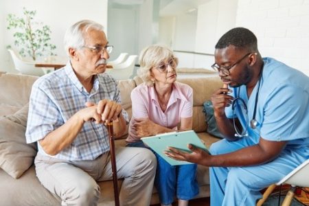 A healthcare professional in scrubs shows a clipboard to an older man and woman sitting on a couch, discussing medical information.