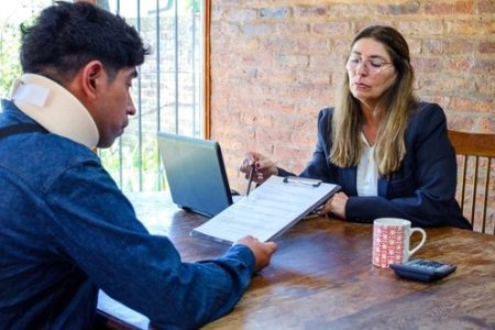 A man wearing a neck brace reviews documents with a woman at a desk; a laptop, mug, and calculator are also on the table.